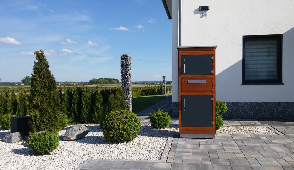 Mypakektasten Referenzfoto Paketkasten aus Holz vom Deggendorfer start-up Mypaketkasten. Paketkasten vor Haus neben Garten mit Büschen und Steinen.
